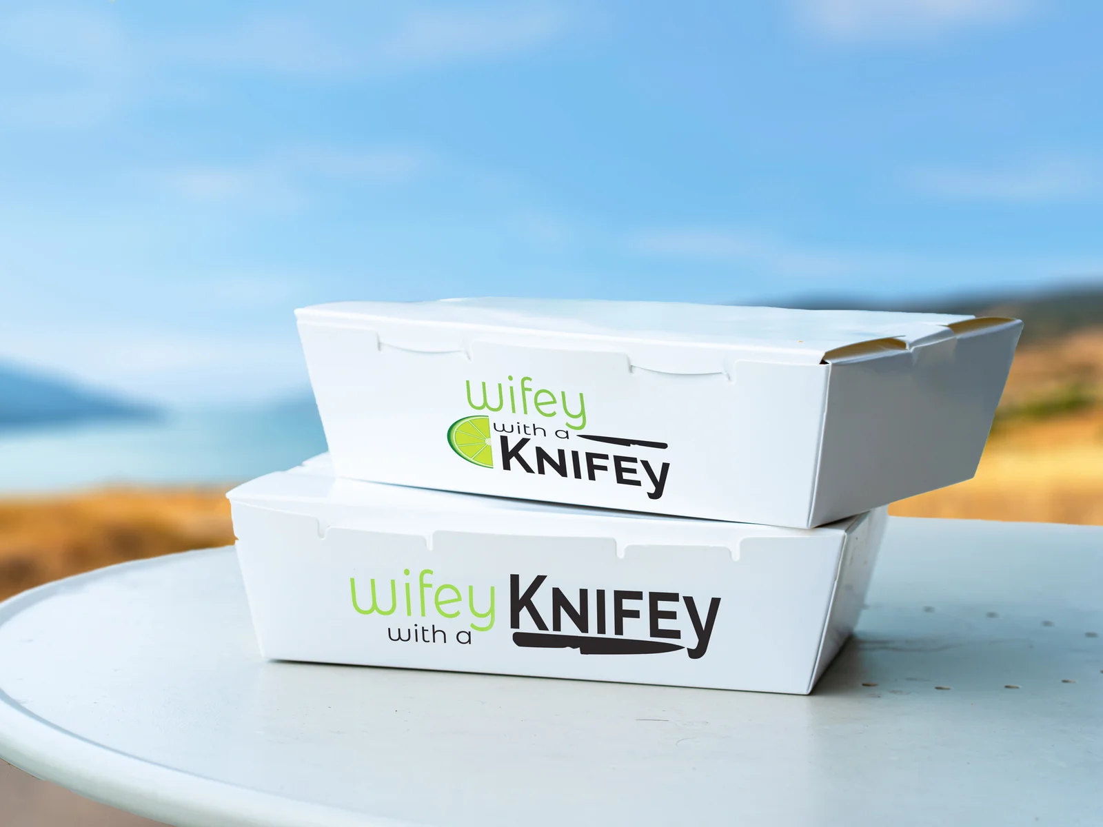 Two white takeout boxes printed with the Wifey with a Knifey wordmark, stacked on a café table above a blurred lake