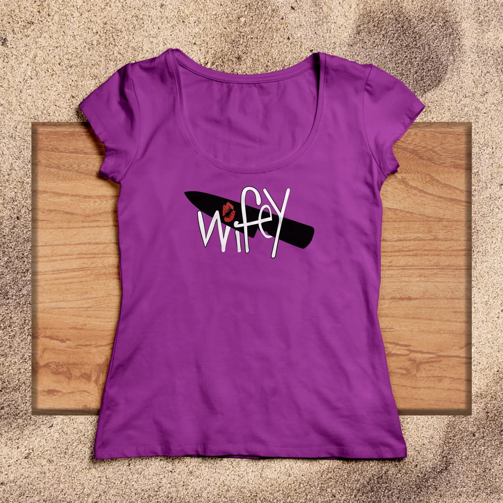 Purple scoop-neck t-shirt printed with the Wifey submark, laid on a wooden board in the sand